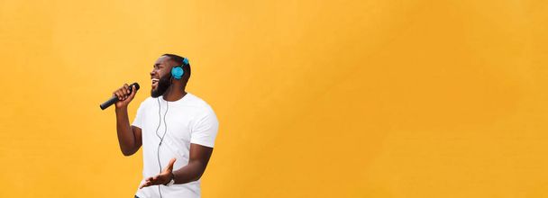 Porträt eines fröhlich positiven Chic. schöner afrikanischer Mann mit Mikrofon und Kopfhörer auf dem Kopf, der Musik lauscht, die Lieder singt und den Wochenendurlaub isoliert auf gelbem Hintergrund genießt. - Foto, Bild