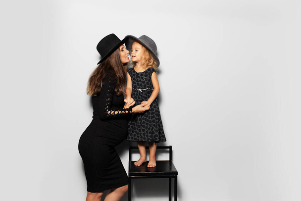 Studio portrait of pregnant smiling woman and child girl looking each ever, wearing hats on white background. Mother and daughter in black. - Photo, Image