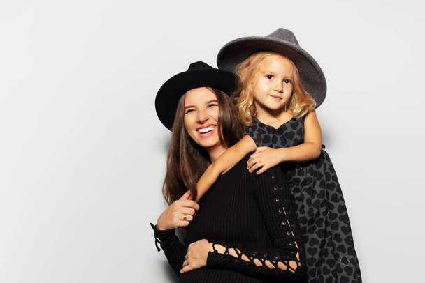 Studio portret van zwangere gelukkige vrouw en kind meisje, het dragen van hoeden op witte achtergrond. Moeder en dochter in het zwart. - Foto, afbeelding