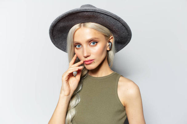 Studio portrait of young blonde attractive girl with earbuds in ears on white background, wearing grey hat. - Photo, Image