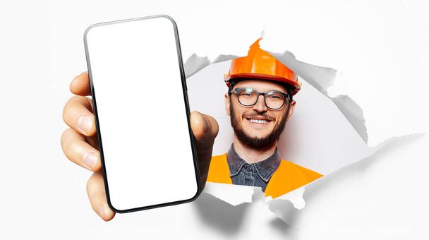 Studio portret młodego uśmiechniętego człowieka, robotnika budowlanego, pokazując duży smartfon z pustym ekranem przez dziurę w papierze. Białe tło. Noszenie okularów i sprzętu bezpieczeństwa. - Zdjęcie, obraz