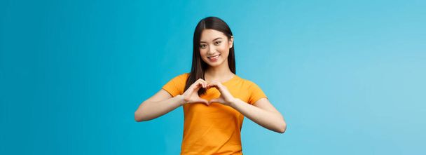 Lovely caring attractive asian girlfriend show heart sign cherish and value relationship, smiling broadly supportive, stand blue background wear yellow t-shirt, promote peace and happiness. - Valokuva, kuva