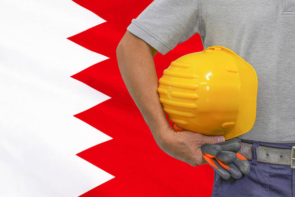 Κοντινό πλάνο του hard hat holding από τον οικοδόμο στο φόντο της σημαίας του Μπαχρέιν. Χέρι εργάτη με κίτρινο σκληρό καπέλο και γάντια. Έννοια των εργαζομένων στη βιομηχανία, τις κατασκευές και τη βιομηχανία στο Μπαχρέιν - Φωτογραφία, εικόνα