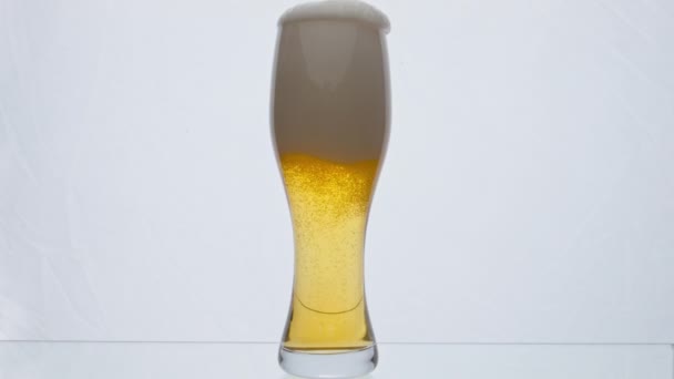 Helder glas vol schuimig smakelijk bier overlopen van de rand in super slow motion close-up. Licht borrelend schuim dat uit transparante beker op een witte ondergrond stroomt. Alcoholdrankconcept - Video
