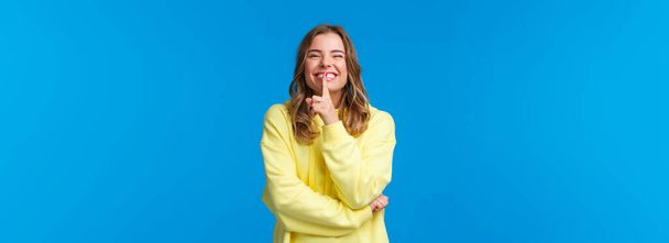 Happy cheerful silly blond caucasian woman in yellow hoodie hiding secret, prepare b-day surprise for boyfriend, shush with index finger pressed to lips as smiling joyfully, blue background. - Valokuva, kuva