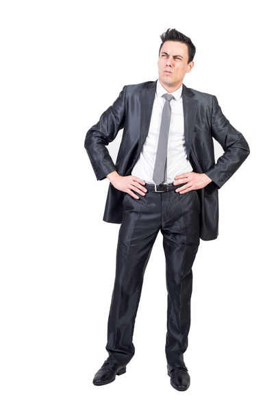 Full body of comic male entrepreneur in suit expressing doubt and looking up while standing with hands on waist against white background - Fotó, kép