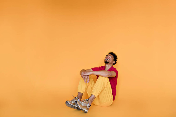 Повноцінне фото стильного темношкірого чоловіка з темним волоссям у яскравому вбранні, що сидить на підлозі і позує на камеру, дивлячись вгору з щасливою посмішкою
.  - Фото, зображення