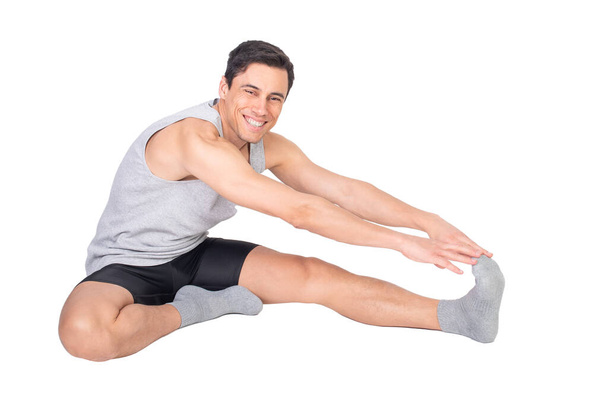 Corpo inteiro de macho esportivo positivo em sportswear olhando para a câmera com sorriso ao esticar o músculo femoral isolado no fundo branco - Foto, Imagem