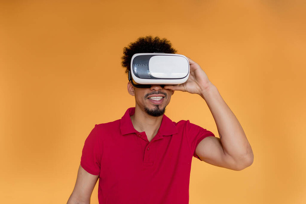 Закрыть фотографию темнокожего мужчины с волнистыми темными волосами, носящего красную футболку в очках VR, играющего в видеоигры на жёлтом фоне  - Фото, изображение