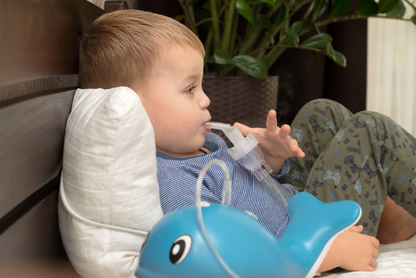 Ein kleiner kaukasischer Junge im gestreiften Pullover liegt im Bett und atmet Hustenmittel mit einem Nibulizer ein. Gesundheitskonzept. Nahaufnahme. Weicher Fokus. - Foto, Bild