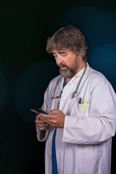 bearded mature doctor dressed in a blue surgical suit and a white coat looking at the mobile phone distractedly thinking about answering healthcare medical professions - Photo, Image