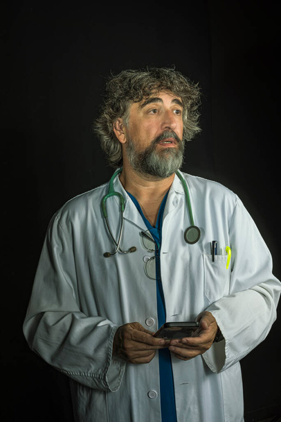 brodaty, dojrzały lekarz ubrany w niebieski garnitur chirurgiczny i biały płaszcz patrzący na telefon komórkowy rozpraszająco myślący o odpowiadaniu na zawody medyczne - Zdjęcie, obraz