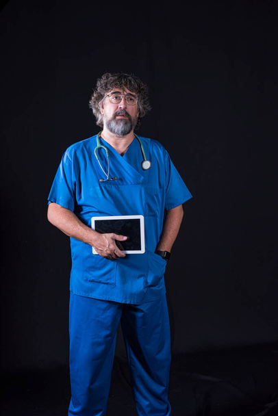 Dojrzały lekarz z brodą ubrany w niebieski strój sali operacyjnej na czarnym tle trzymający tablet patrzący na kamery medyczne zawody medyczne - Zdjęcie, obraz