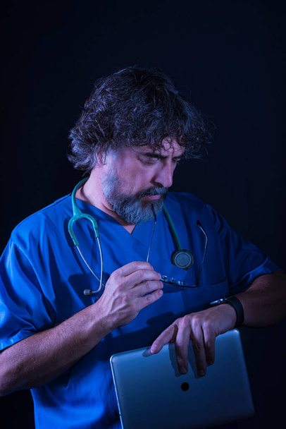 mature doctor with a beard dressed in a blue operating room suit on a black background consulted the time healthcare medical professions - Photo, Image