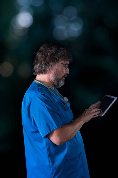 mature doctor with a beard dressed in a blue operating room suit on a black background consulting a tablet healthcare medical professions - Photo, Image