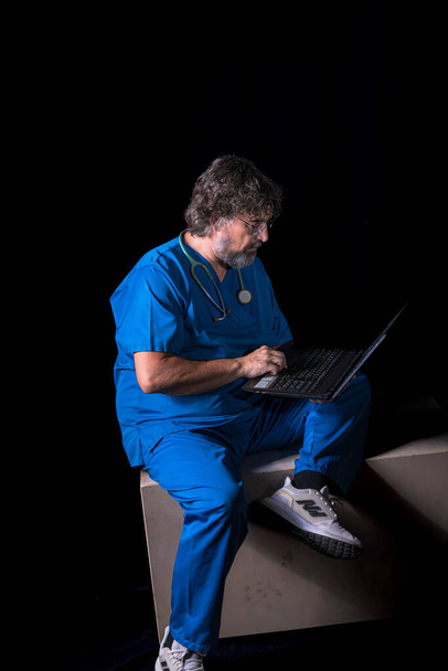 bearded mature doctor dressed in blue operating room suit sitting on a wooden box consulting a computer healthcare medical professions - Fotoğraf, Görsel