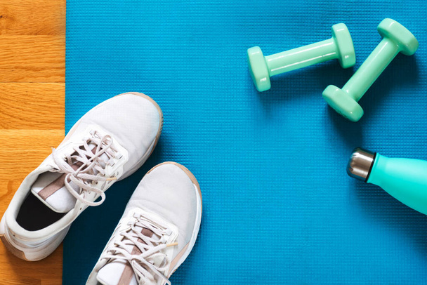 Light green dumbbells, bottle with water and white sneakers on blue sports mat background. Training at home concept. Top view, flat lay, copy space. - Photo, Image