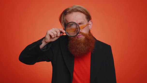 Badacz naukowiec trzymający szkło powiększające blisko twarzy, patrzący w kamerę z dużym zoomem śmiesznych oczu, szukający, analizujący. młody ruda facet odizolowany na pomarańczowy studio tło - Materiał filmowy, wideo