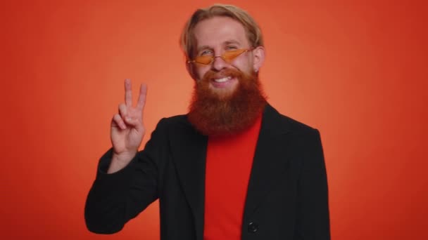 Bearded man in black jacket showing victory sign, hoping for success and win, doing peace gesture, smiling with kind optimistic expression. Young redhead guy boy isolated on orange studio background - Footage, Video