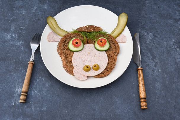 Sandwich en forme de portrait d'un taureau joyeux fait de pain noir, de saucisses et de légumes sur une assiette, sur un fond sombre avec un décor de thuja et de branches de houx. Photo de haute qualité - Photo, image