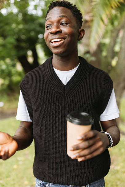 Молодой афроамериканец улыбается с чашкой кофе на улице. Молодой черный студент на кофе-брейке в парке - Фото, изображение