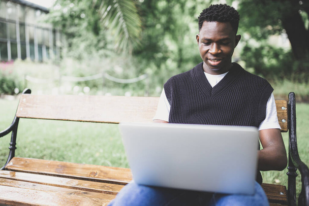 Jonge zwarte man in vest werkt buiten aan laptop computer, kijkt naar het scherm, glimlacht. Studenten zitten op een bank op de universiteitscampus, genieten van freelance afstandsonderwijs - Foto, afbeelding
