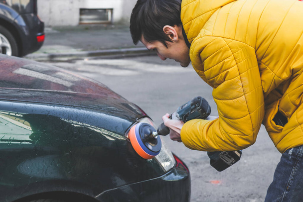 The hands of a young caucasian guy are drilling a drill with a disk with a sponge and polishing the headlight of his car with a detergent soap, close-up side view.The concept of a home car wash and cleaning with polishing car headlights. - Foto, Imagem