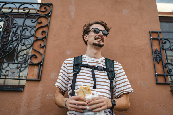 One man young adult modern caucasian male in the city in sunny day stand and eat sandwich fast food concept urban life copy space tourist eating in front of building wall real person portrait - Photo, Image