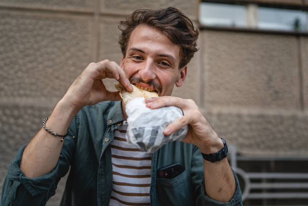 Genç, yetişkin, beyaz bir erkek, güneşli bir günde şehirde duruyor ve sandviç yiyor. Fast food konsepti. Şehir hayatı. - Fotoğraf, Görsel