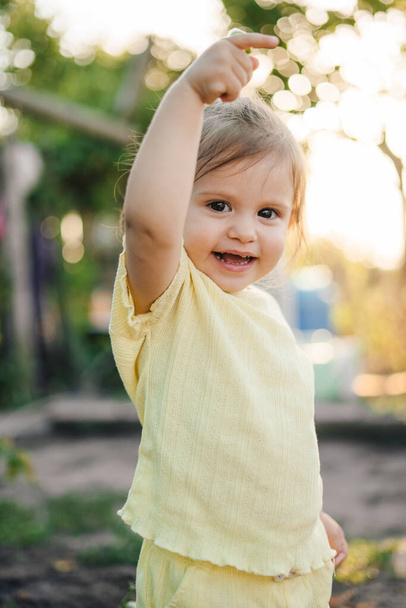 Porträt eines kleinen Mädchens, das glücklich mit dem Finger auf die rechte Hand zeigt. Grünes Porträt. Babyentwicklung. Sommerliche Natur. Grüne Natur. - Foto, Bild