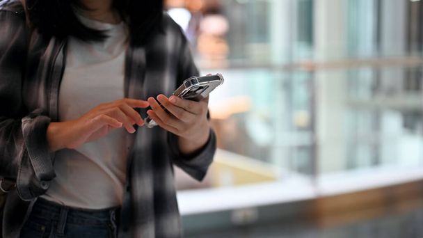 A hipster young Asian female in a flannel shirt uses her smartphone to message her friends while waiting in the shopping mall. cropped image - Фото, зображення