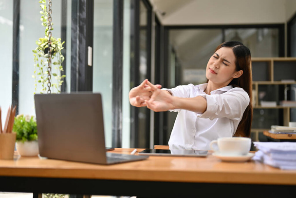 Zmęczona i niewygodna młoda azjatycka bizneswoman lub pracownica biurowa przy biurku, wyciągająca ręce, cierpiąca na syndrom biurowy. przepracowana koncepcja przedsiębiorców - Zdjęcie, obraz