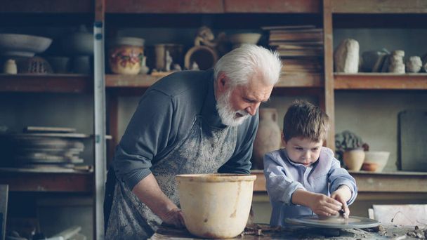 Pottery teacher senior adult in muddy apron is helping young student to form pot from piece of clay on throwing-wheel. Cute boy is awkward, and craftsman is helping him. - 写真・画像