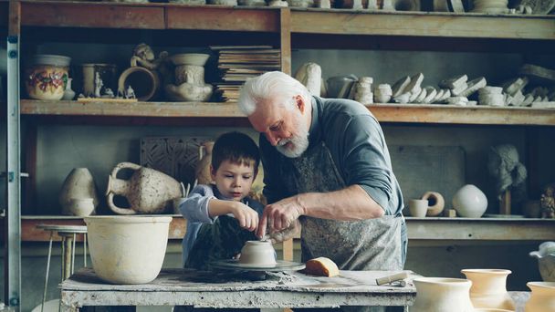 Caring senior grandfather is showing young cute grandson how to work with clay on throwing-wheel in small workshop. Pottery, family hobby and handicraft concept. - Photo, image