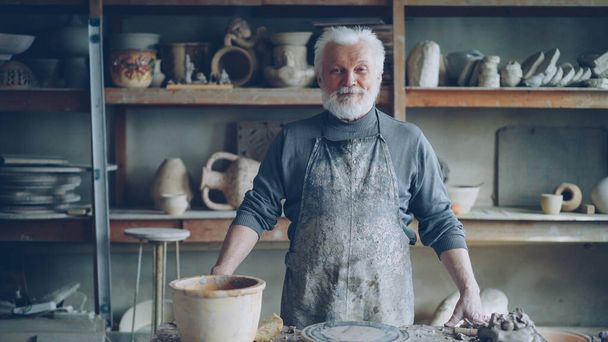 Portrait of senior silver-haired potter in muddy apron standing alone at table in workshop and looking at camera. Shelves with handmade vases and pots in background. - Photo, image