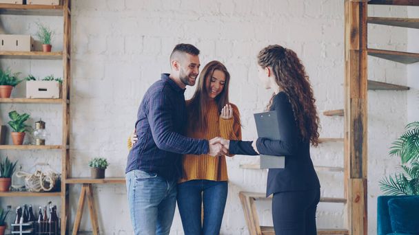 Friendly realtor is giving keys to young couple buyers of new house, happy spouses are hugging , smiling man is shaking hands with broker making deal. - Photo, image
