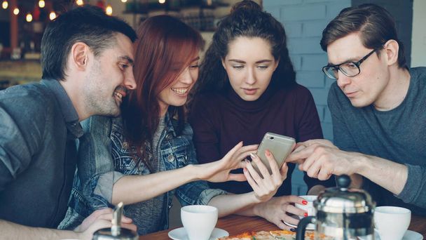 Cheerful young men and women close friends are using smartphone and talking while having lunch in nice cozy cafe. Dining, modern technology and friendship concept. - 写真・画像