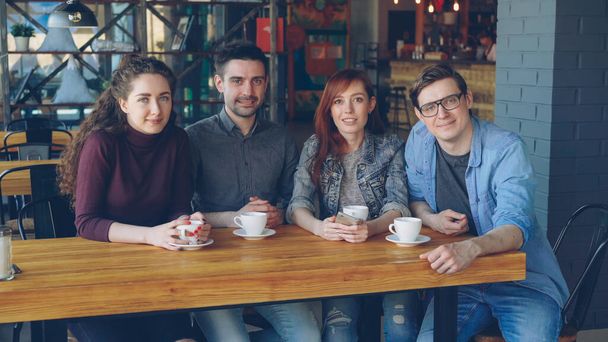 Portrait of four smiling young people friends in casual clothes sitting at table in spacious cafe with tea cups and looking at camera. Eating out and friendship concept. - Fotoğraf, Görsel