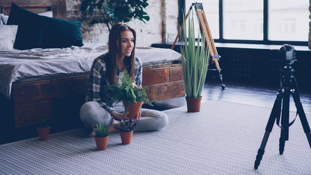 Young woman famous blogger is recording video about pot flowers using camera while sitting on bedroom floor near double bed. Girl is talking and showing green plants. - Foto, afbeelding