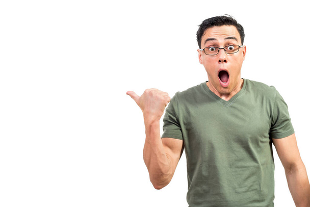 Verblüffter männlicher Nerd in T-Shirt und Brille blickt mit geöffnetem Mund in die Kamera und zeigt vor weißem Hintergrund mit dem Daumen weg - Foto, Bild