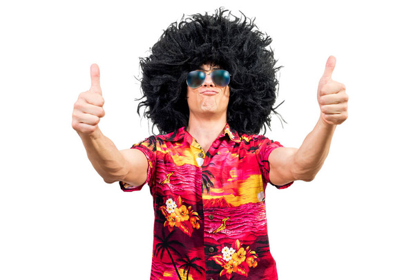 Positiver Mann im lebhaften Hemd mit Sonnenbrille und Afro-Perücke, schmollende Lippen und gestikulierende Daumen vor weißem Hintergrund - Foto, Bild