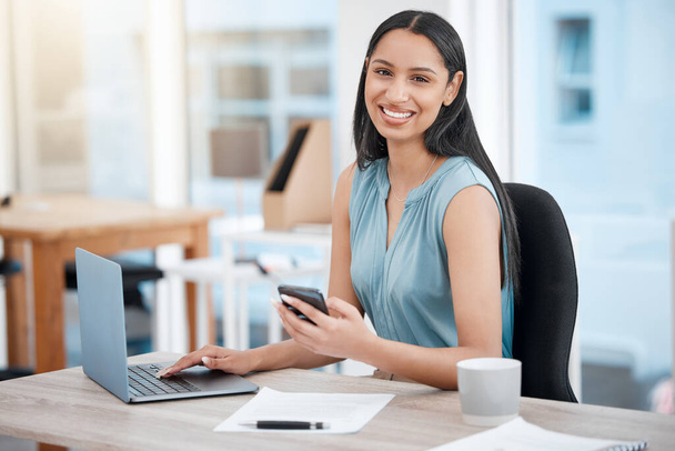 Confident young mixed race businesswoman texting on cellphone while working on laptop in an office. One female only checking browsing apps and online media on smartphone to manage her startup plans. - Photo, Image