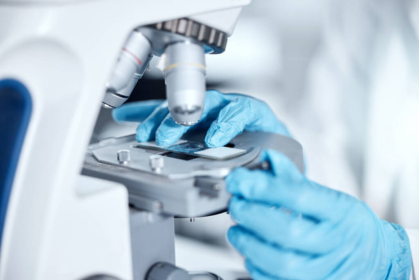 Closeup of the hands of one young woman wearing a labcoat and gloves and looking at medical samples on a microscope in her lab. A mixed race female scientist wearing goggles conducting research. - Foto, Imagen