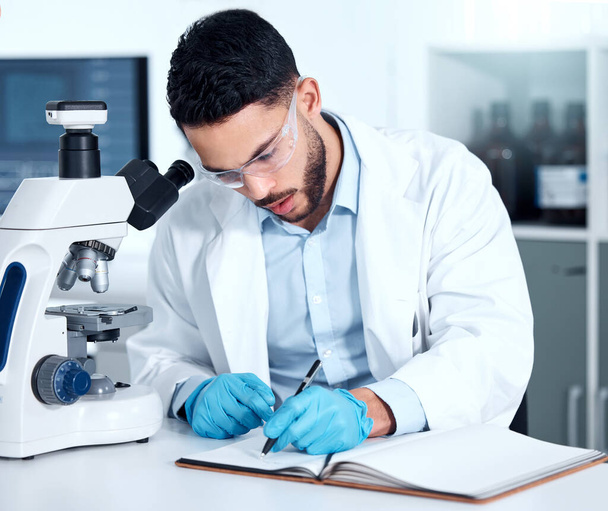 Serious young mixed race male scientist wearing glasses and gloves while writing in a notebook sitting at a desk alone working in a lab. One hispanic lab worker wearing goggles and taking notes. - Foto, afbeelding