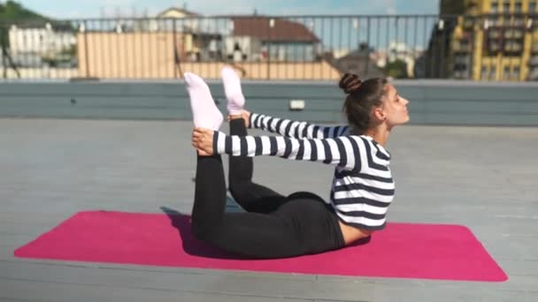 Young sporty happy woman in sportswear doing stretching exercises on yoga mat on house roof in early morning - Filmati, video