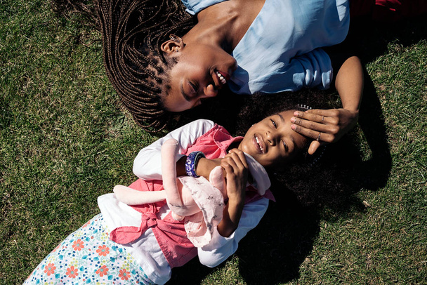Urocza afro-dziewczyna przytulająca pluszowego zwierzaka leżącego na trawie ze swoją matką. Uśmiechają się i dobrze się bawią. - Zdjęcie, obraz