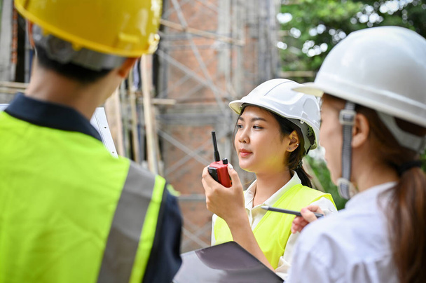 A professional young Asian female engineer using a walkie talkie to communicate with her worker in the construction site. - Fotoğraf, Görsel
