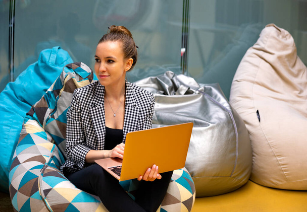 A young girl is sitting on a soft pouf with a laptop, working. She was distracted by the interlocutor, smiling. High quality photo - Fotografie, Obrázek