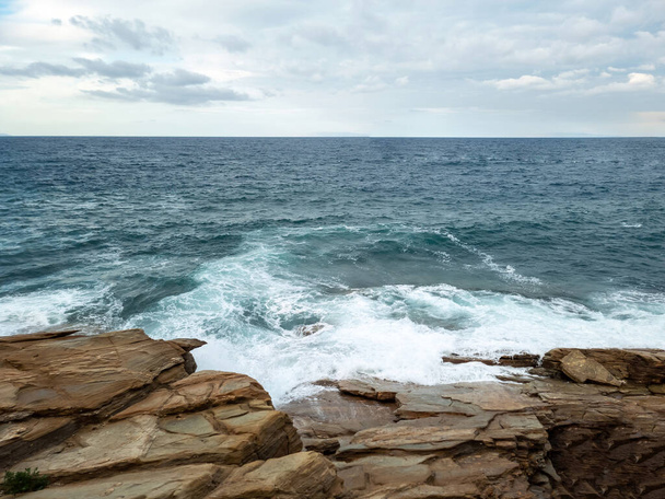 Islak kaya yüzeyinde dalgalı su köpüğü, bulutlu gökyüzü arkaplanı. Boş kayalık plaj, Yunan adası Cyclades, Yunanistan. Yaz tatili şablonu, boşluk. - Fotoğraf, Görsel