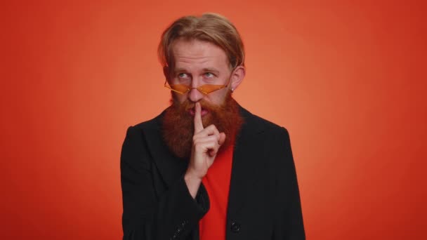 Shh be quiet please. Portrait of bearded redhead man 20 years old presses index finger to lips makes silence gesture sign do not tells secret. Young adult handsome guy on orange studio background - Footage, Video